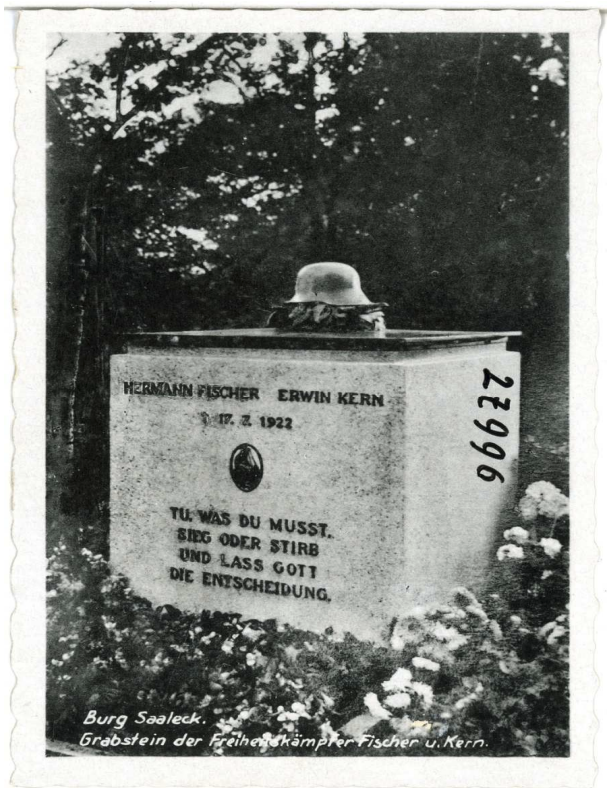 Grabstein mit aufgesetztem Stahlhelm, eingravener Inschrift und Blumen vor Bäumen